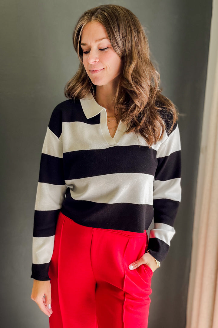 A woman wearing a black and off-white striped long-sleeve knit polo sweater and bright red trousers, standing against a dark gray wall.