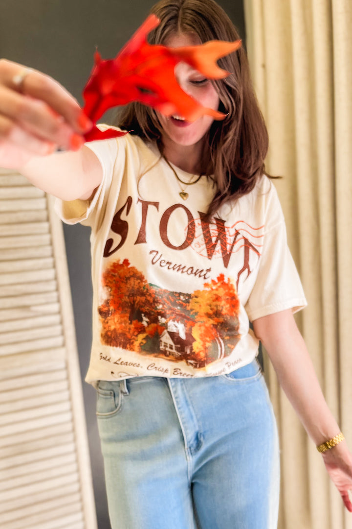 A woman wearing a cream-colored graphic t-shirt with 'STOWE Vermont' and an autumn leaf print, paired with light blue jeans, holding a red leaf in front of her face.