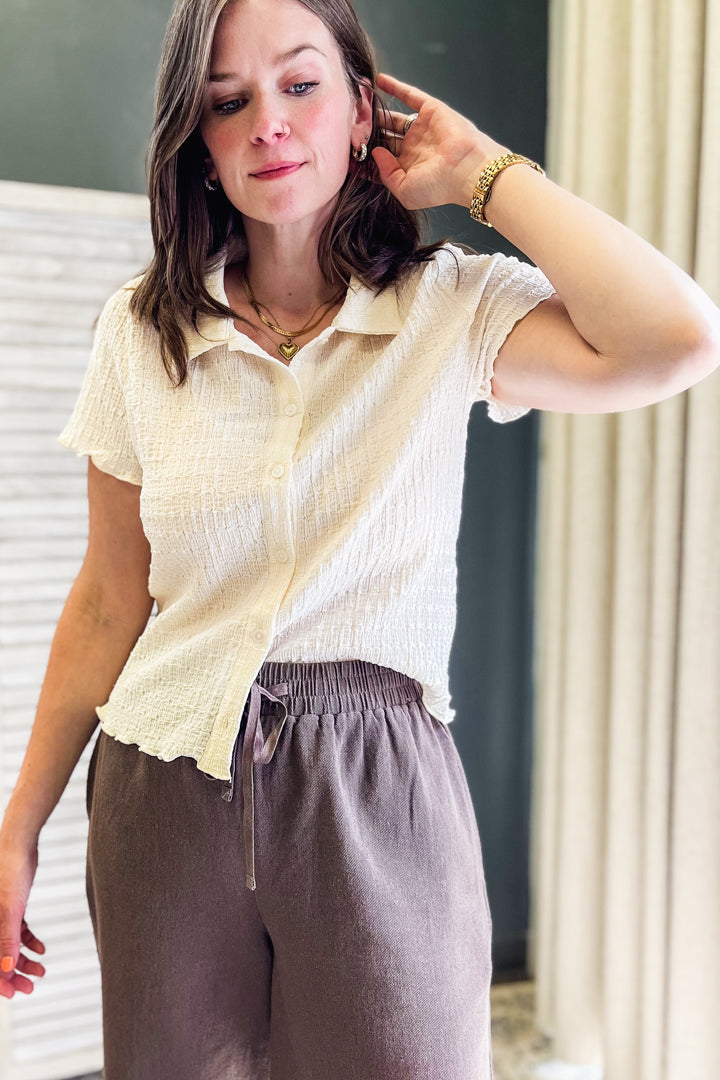 A woman wearing a cream-colored short-sleeve textured button-up shirt and brown drawstring trousers, standing indoors.