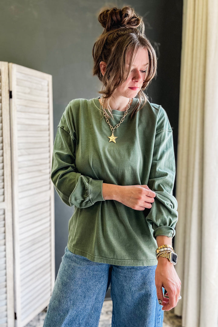 A woman wearing an oversized sage green long-sleeve t-shirt and blue jeans, accessorized with gold necklaces and bracelets, standing indoors next to a white folding screen.