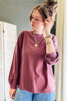 A woman wearing a washed mauve long-sleeve t-shirt and blue jeans, accessorized with a gold star pendant necklace and bracelets, standing indoors next to a white folding screen.