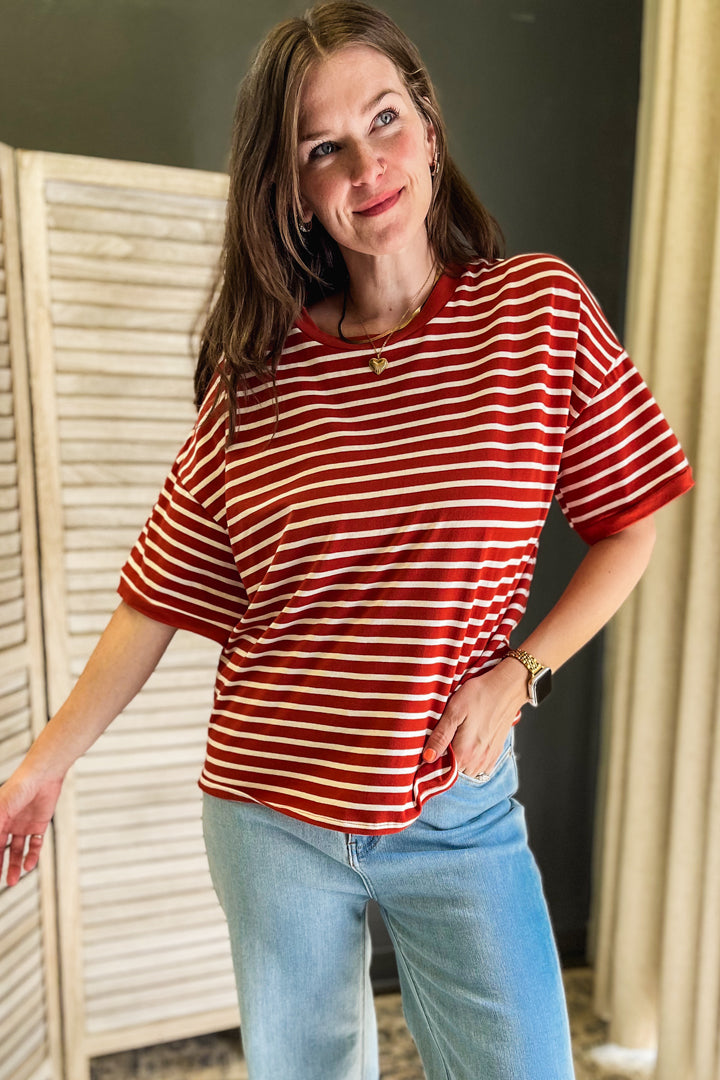 A woman wearing a rust and white horizontally striped short-sleeve t-shirt and light blue jeans, standing indoors in front of a white folding screen.