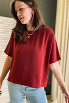 A woman wearing a short-sleeve, relaxed-fit top in a deep rust color, paired with light blue jeans. She is wearing layered gold necklaces and a gold bracelet, standing indoors against a background with neutral-toned curtains and a dark wall.