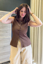 A woman wearing a brown sleeveless button-front knit vest and beige cargo pants, standing indoors with her hands in her hair.