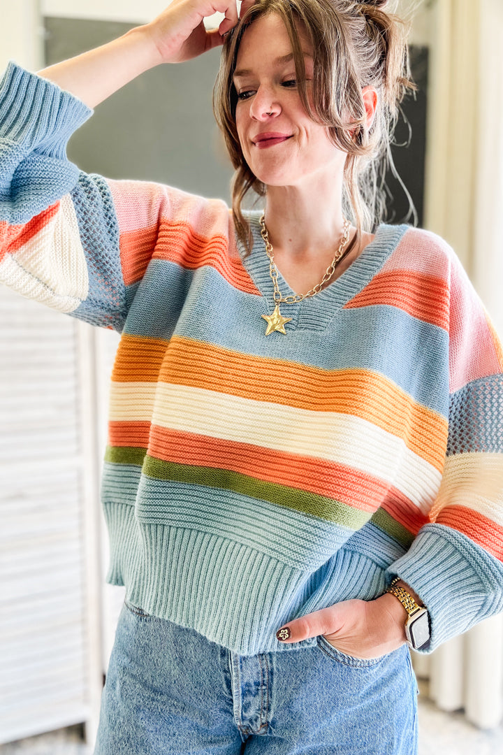 A woman wearing a pastel striped knit sweater with horizontal bands of light blue, orange, cream, and olive green, paired with blue jeans.