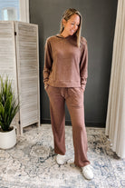 Woman wearing a brown outfit standing in a room with a plant and curtains.