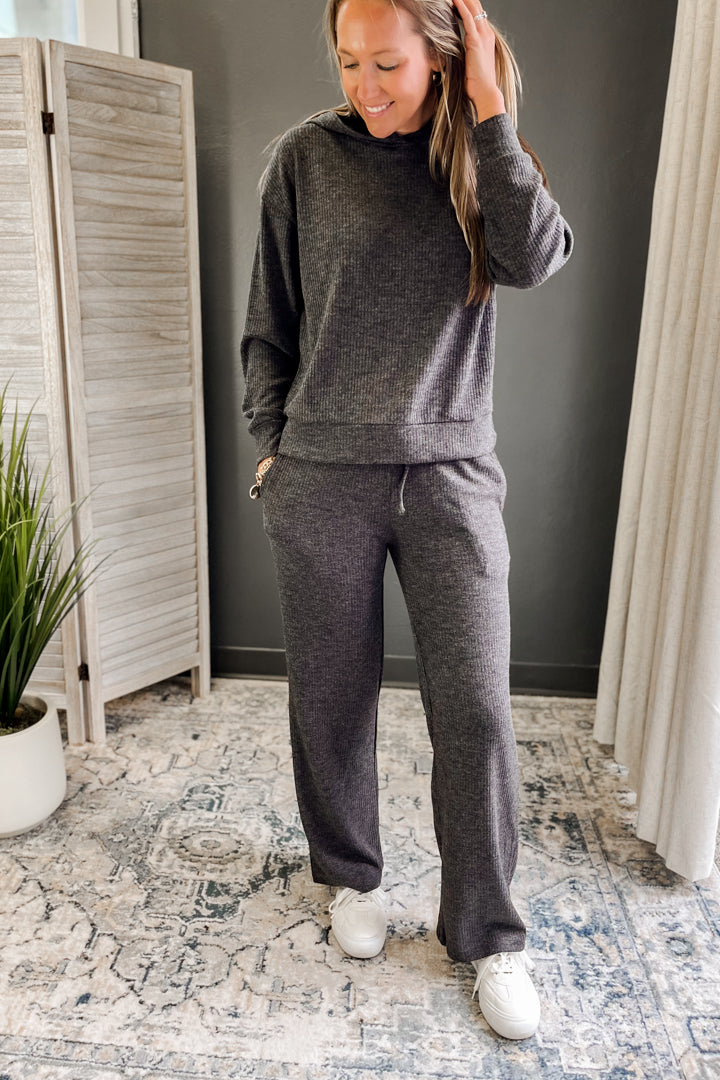 Woman wearing a gray tracksuit standing in a room with a plant and curtains.