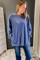 Woman wearing a blue sweater and jeans in a room with a dark wall and light-colored curtains.