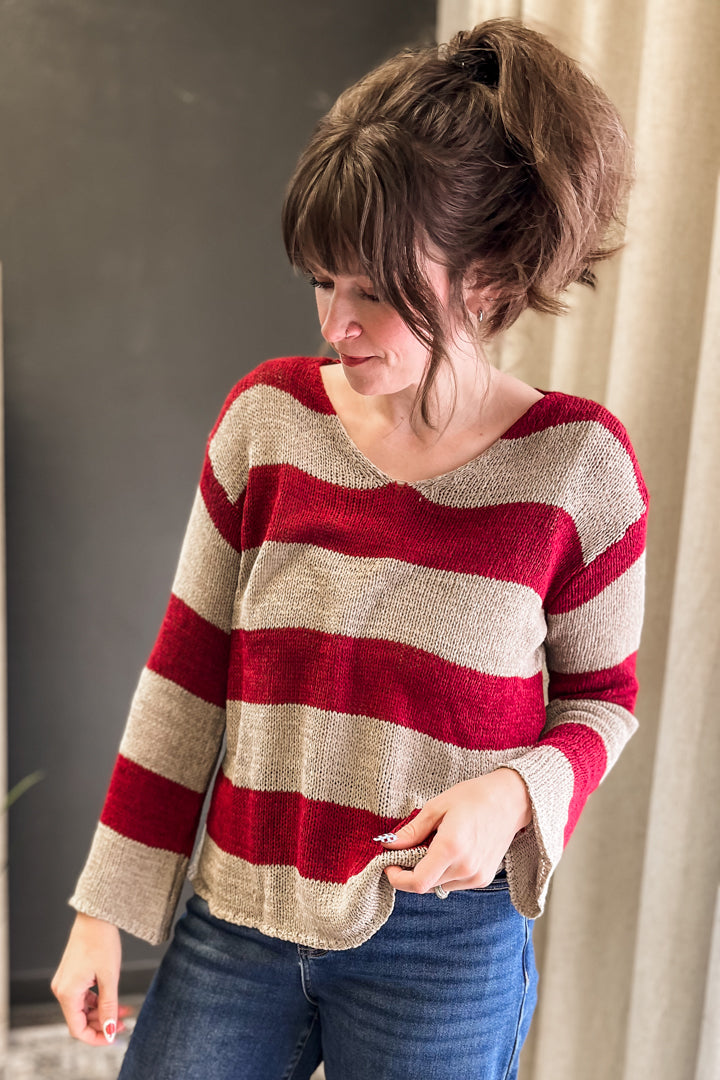 A woman wearing a red and beige striped knit sweater with blue jeans, standing indoors against a dark gray wall and beige curtain background.