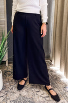 A person wearing high-waisted, wide-leg navy trousers with a white textured knit top and black flat shoes, standing on a patterned rug in an indoor setting.