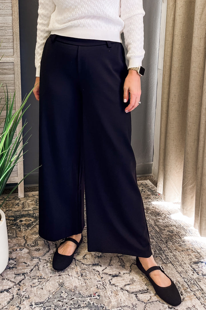 A person wearing high-waisted, wide-leg navy trousers with a white textured knit top and black flat shoes, standing on a patterned rug in an indoor setting.