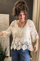 A woman wearing a cream-colored, semi-sheer crochet lace top with three-quarter sleeves and a tie-front detail, paired with blue jeans.