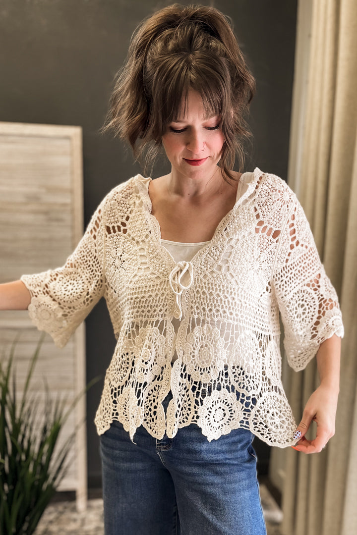 A woman wearing a cream-colored, semi-sheer crochet lace top with three-quarter sleeves and a tie-front detail, paired with blue jeans.