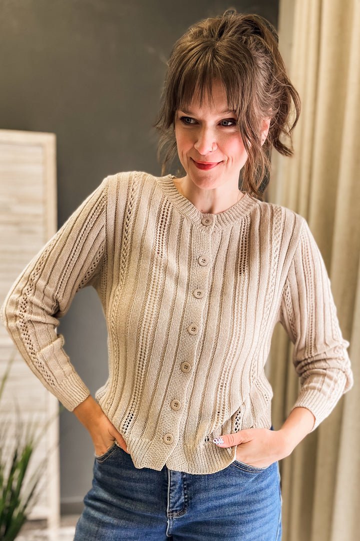 A woman wearing a beige cable-knit button-up cardigan sweater with blue jeans, standing indoors with her hands on her hips.