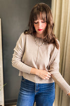 A woman wearing a beige ribbed knit cardigan sweater and blue jeans, standing indoors against a dark gray wall and beige curtain.