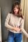 A woman wearing a beige ribbed knit cardigan sweater and blue jeans, standing indoors against a dark gray wall and beige curtain.