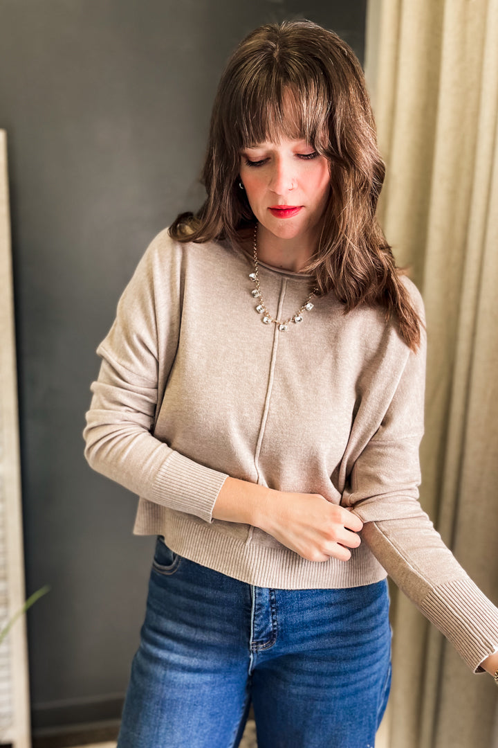 A woman wearing a beige ribbed knit cardigan sweater and blue jeans, standing indoors against a dark gray wall and beige curtain.