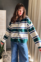 A woman wearing a green, white, and navy blue striped knit cardigan sweater with blue jeans, standing in an indoor setting with curtains and a plant visible.
