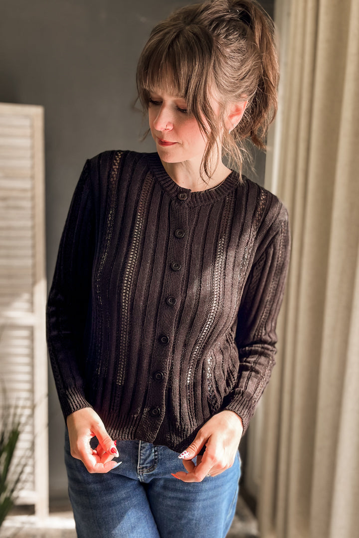 A woman wearing a dark brown ribbed button-front cardigan sweater and blue jeans, standing indoors near a window with curtains.