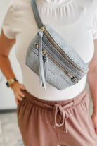 Person wearing a gray crossbody bag with gold zippers, white top, and brown pants.