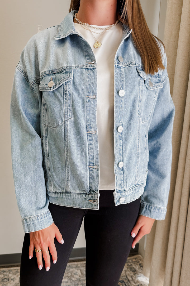 Person wearing a light blue denim jacket over a white shirt and black pants indoors.