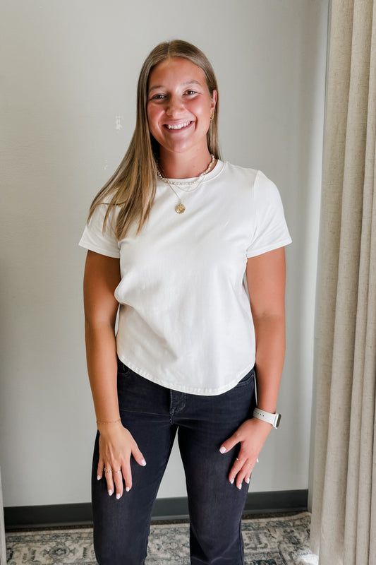 Woman wearing a white t-shirt and dark pants standing indoors against a neutral wall.