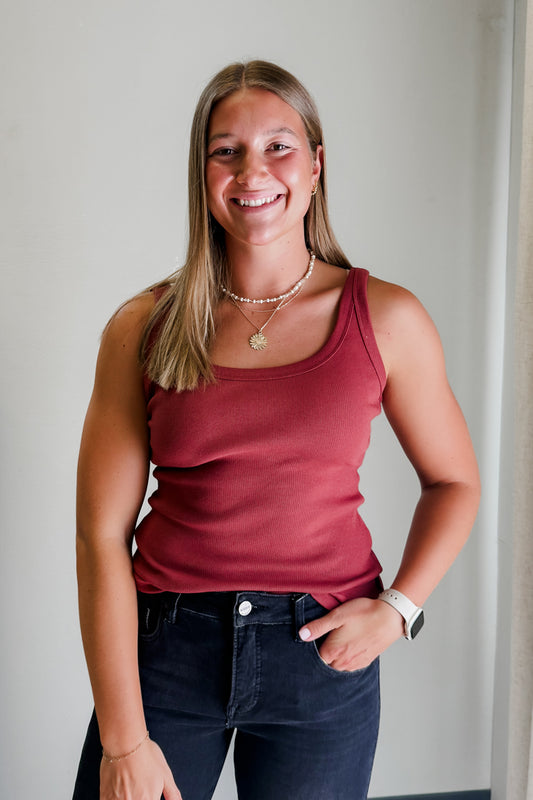 Woman wearing a red tank top and dark jeans against a plain background