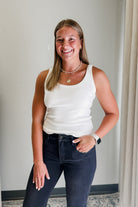 Woman wearing a white tank top and dark jeans standing against a plain wall.