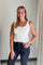 Woman wearing a white tank top and dark jeans standing against a plain wall.