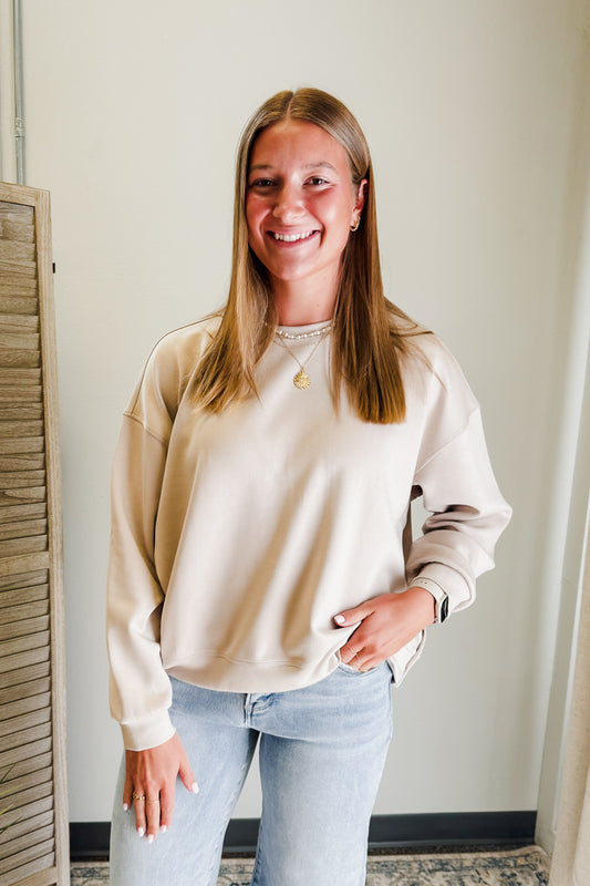 Woman wearing a beige sweatshirt and light blue jeans standing against a white wall.
