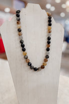 A beaded necklace made of polished round gemstone beads in black, brown, gray, and translucent amber tones, displayed on a white jewelry bust.