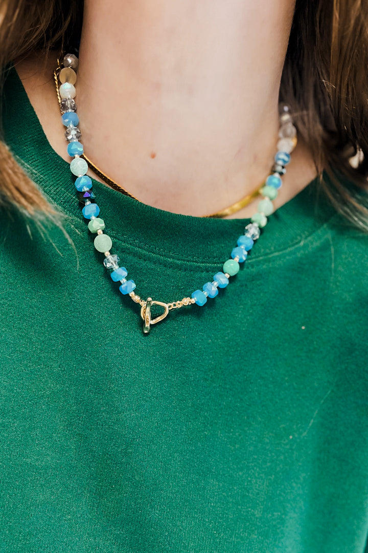 A close-up of a person wearing a beaded necklace with light blue, mint green, and white beads, a gold heart-shaped clasp, and a thin gold chain layered underneath, paired with a green sweatshirt.