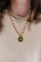 A close-up of a person wearing a gold chain necklace with a cushion-cut green gemstone pendant, paired with a beige crewneck sweatshirt.
