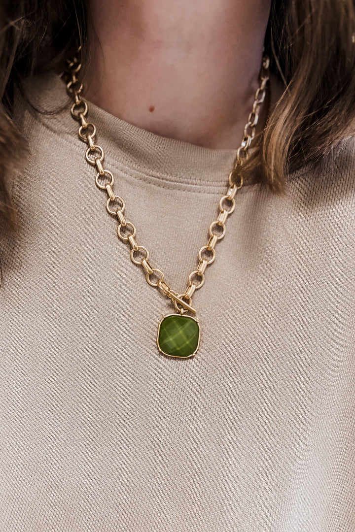 A close-up of a person wearing a gold chain necklace with a cushion-cut green gemstone pendant, paired with a beige crewneck sweatshirt.