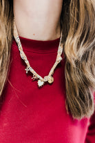 A close-up of a person wearing a gold-toned charm necklace over a red knit top. The necklace features a patterned chain with small dangling charms, including a bow, a flower, and a round charm.