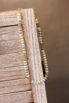 A beaded necklace draped over a rustic wooden surface, featuring small round beads in pale yellow, light blue, white, and muted lavender on a gold-tone chain.