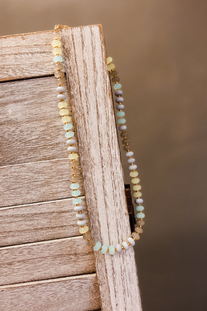A beaded necklace draped over a rustic wooden surface, featuring small round beads in pale yellow, light blue, white, and muted lavender on a gold-tone chain.