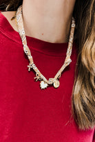 A close-up of a person wearing a gold-toned charm necklace over a red crewneck top. The necklace features a patterned chain with various charms including a starfish, a small cross, a heart, and a coin pendant.