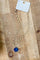 A gold chain necklace with a blue cushion-cut stone pendant, attached to a white paper display card, laid flat on a wooden surface.