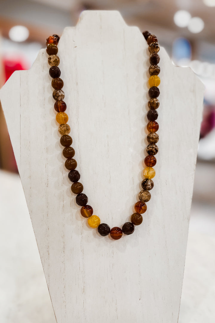 A beaded necklace made of polished round gemstone beads in earthy brown, amber orange, and translucent yellow tones, displayed on a white jewelry bust.