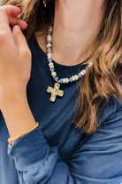 A close-up lifestyle shot of a person wearing a beaded necklace with a gold cross pendant and a matching gold beaded bracelet, paired with a navy blue long-sleeve shirt.