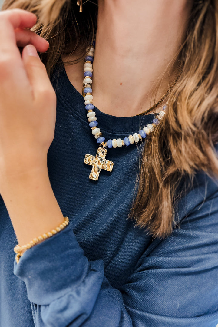 A close-up lifestyle shot of a person wearing a beaded necklace with a gold cross pendant and a matching gold beaded bracelet, paired with a navy blue long-sleeve shirt.