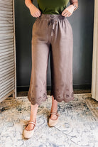 A person wearing loose, wide-leg cropped pants in a muted taupe-brown color with floral eyelet embroidery along the scalloped hems. The pants feature an elastic waistband with a drawstring.