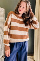 A woman wearing a brown and cream horizontally striped waffle-knit sweater and blue wide-leg pants, standing in an indoor setting with a dark green wall and curtains.