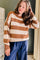 A woman wearing a brown and cream horizontally striped waffle-knit sweater and blue wide-leg pants, standing in an indoor setting with a dark green wall and curtains.