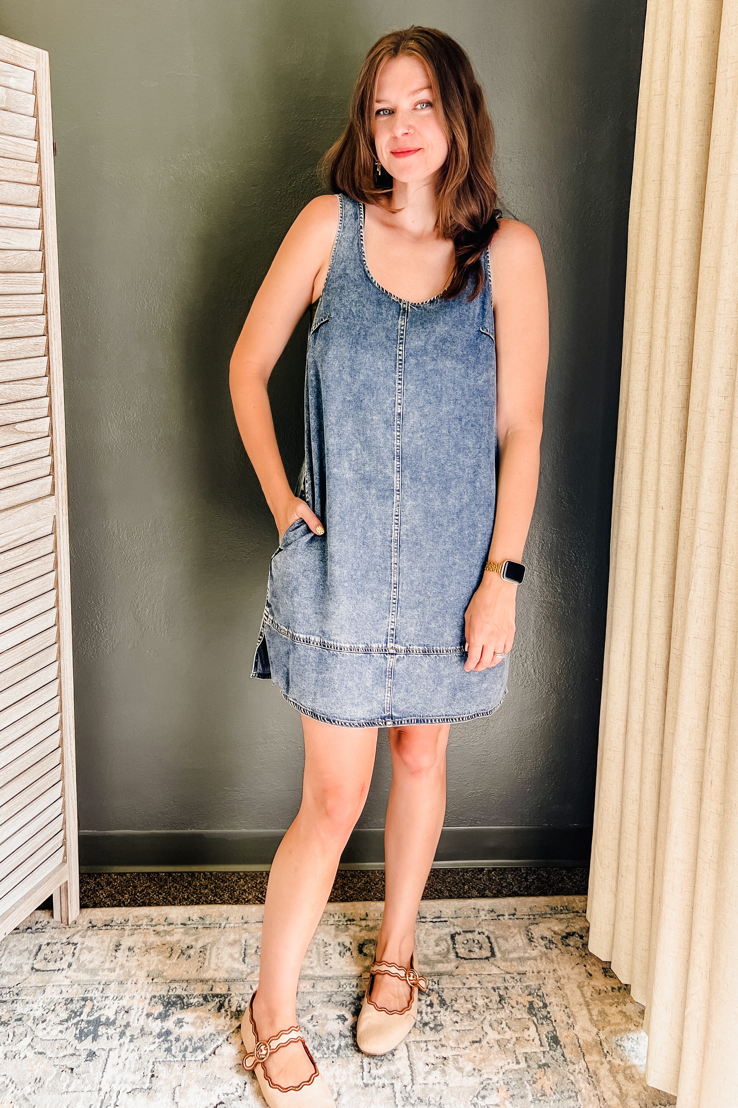 A woman modeling a sleeveless, V-neck denim pinafore dress with side pockets, paired with tan flat sandals, standing in an indoor setting with a dark gray wall, patterned tile floor, and cream curtains.