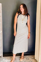 A woman modeling a sleeveless midi dress with horizontal navy and off-white stripes, featuring a buttoned henley neckline and black shoulder panels, standing against a dark gray wall.