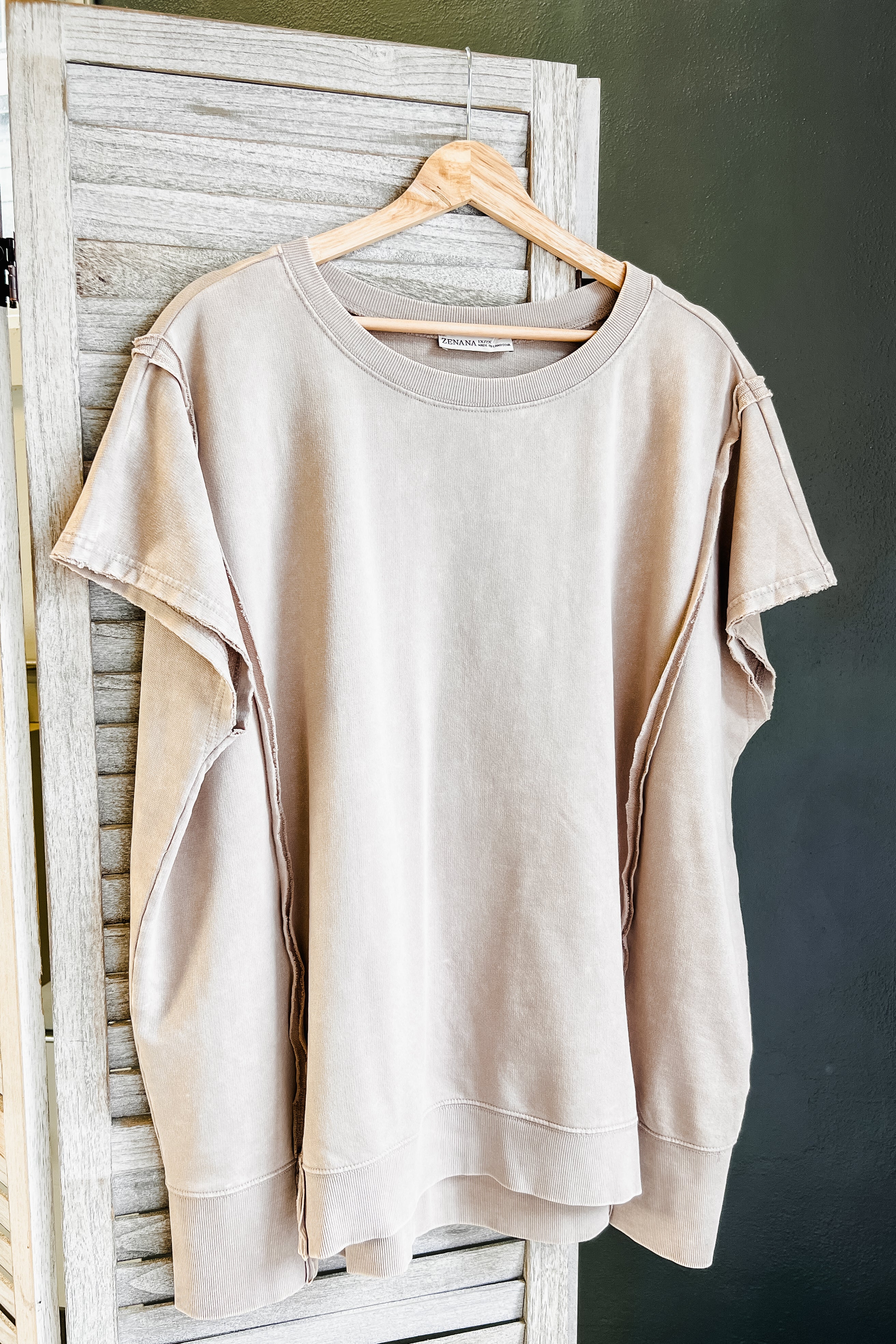 A light beige crewneck sweatshirt with unique draped side slits, hanging on a wooden hanger against a rustic white wooden screen and a dark green wall.