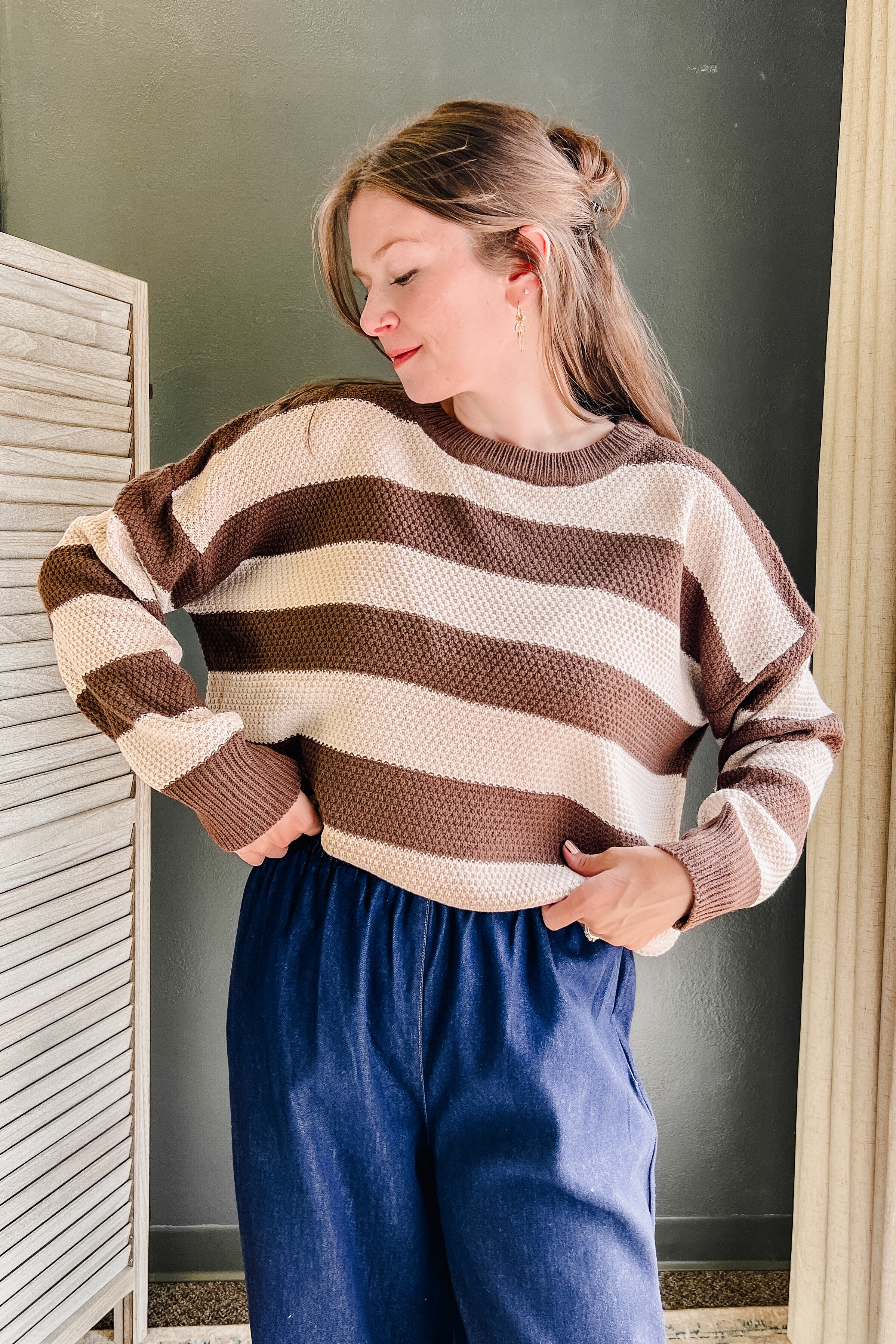 A woman wearing a brown and cream striped knit sweater and blue pants, standing indoors against a dark gray wall.