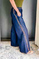 A person wearing wide-leg blue denim pants with  cheetah stripe running down the side, paired with a green top and beige sandals, standing on a patterned rug.